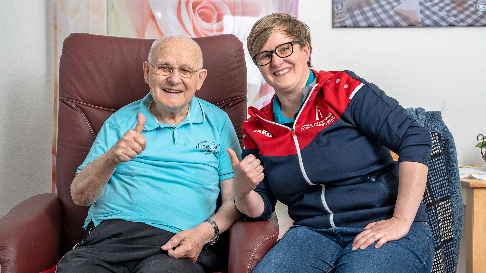 Betreutes Wohnen: Selbstbestimmt, aktiv und sicher im Alter. Mann und Pflegerin lächeln in die Kamera und halten die Daumen hoch.